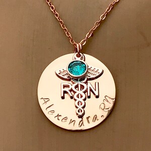 May include: Rose gold necklace with a round pendant featuring a medical caduceus symbol, the letters "RN" and the name "Alexandra RN". A small turquoise gemstone is set above the caduceus.