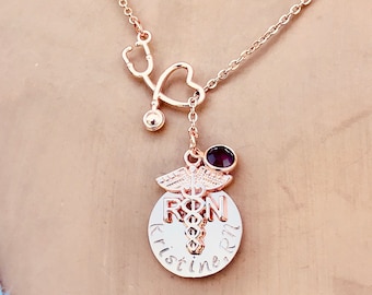 May include: Rose gold necklace with a stethoscope charm, a heart charm, a small purple gemstone, and a round pendant with the words "RN" and "Kristine" engraved on it.