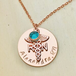 May include: Rose gold necklace with a round pendant featuring a medical caduceus symbol, the letters "RN", and the name "Alexandra RN". A small turquoise gemstone is attached to the top of the pendant.