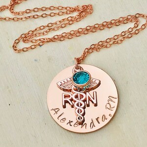 May include: Rose gold necklace with a round pendant featuring a medical caduceus symbol, the letters "RN" and the name "Alexandra RN". A small turquoise gemstone is set in the center of the pendant.