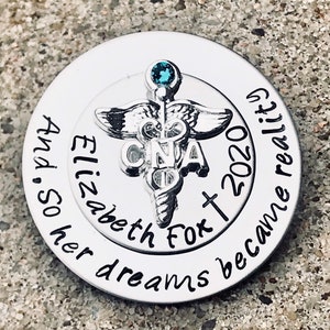 May include: A silver round charm with a caduceus symbol and the text "CNA" in the center. The text "Elizabeth Fox + 2020" is inscribed around the caduceus. The text "And so her dreams became reality" is inscribed around the outer edge of the charm.