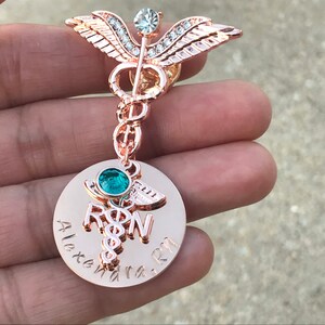May include: Rose gold medical caduceus pin with clear and green rhinestones. The pin has a round charm with the words "RN" and "Alexandra RN" engraved on it.