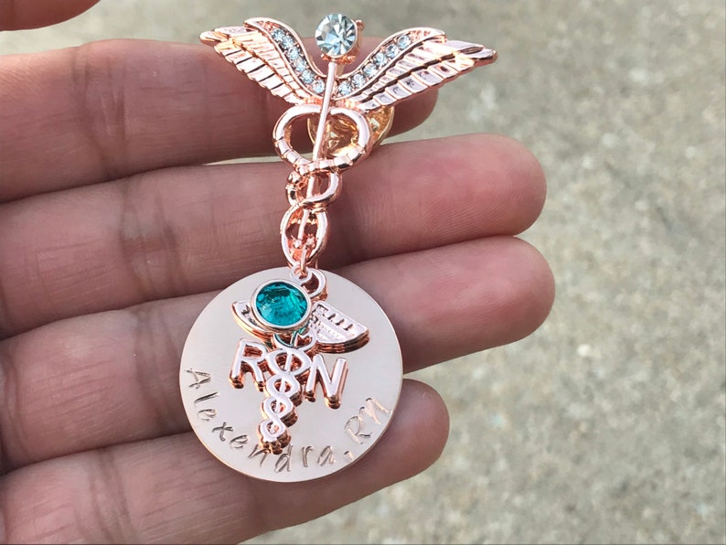 May include: Rose gold medical caduceus pin with clear and green rhinestones. The pin has a round charm with the words "RN" and "Alexandra RN" engraved on it.