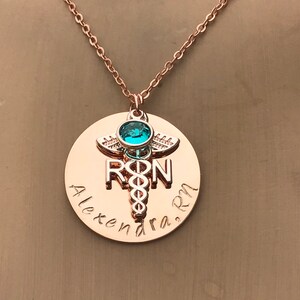 May include: Rose gold necklace with a round pendant featuring a medical caduceus symbol, the letters "RN" and the name "Alexandra RN". A turquoise gemstone is set in the caduceus.