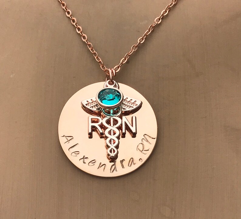 May include: Rose gold necklace with a round pendant featuring a medical caduceus symbol, the letters "RN" and the name "Alexandra RN". A turquoise gemstone is set in the caduceus.