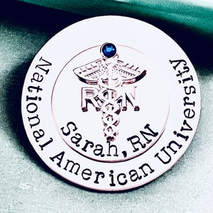 May include: A gold-toned round pin with the words "National American University" and "Sarah, RN" engraved around the edge. A silver medical caduceus symbol with a blue gemstone is in the center.
