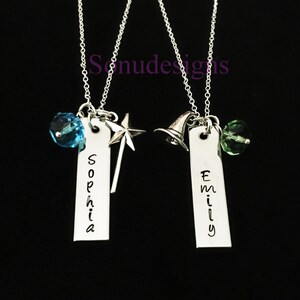 Może przedstawiać: Dwa srebrne naszyjniki z spersonalizowanymi zawieszkami z imionami, jeden z niebieskim kryształem i zawieszką w kształcie gwiazdy, drugi z zielonym kryształem i zawieszką w kształcie kapelusza czarownicy. Zawieszki z imionami to "Sophia" i "Emily".