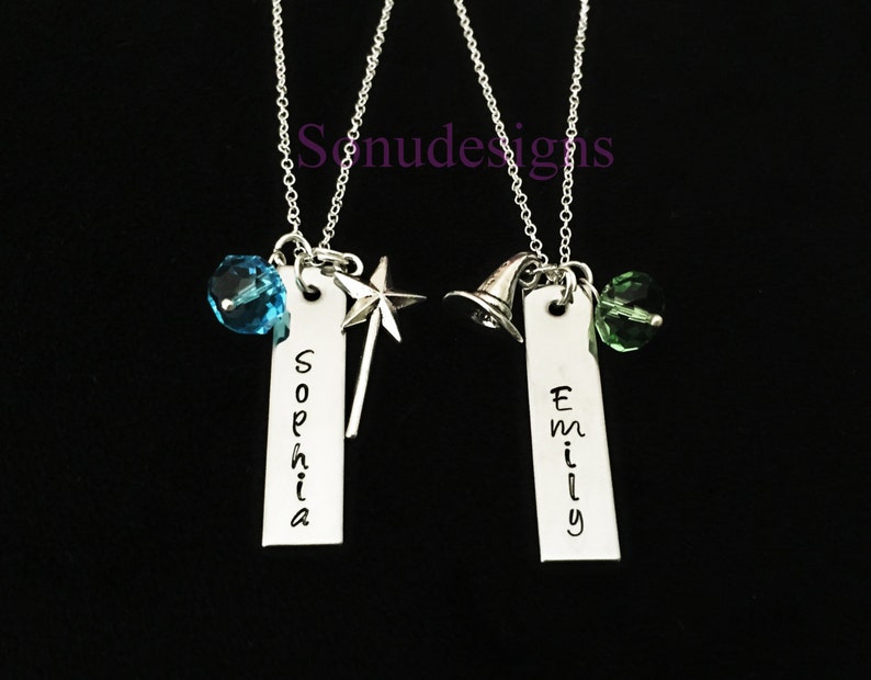Może przedstawiać: Dwa srebrne naszyjniki z spersonalizowanymi zawieszkami z imionami, jeden z niebieskim kryształem i zawieszką w kształcie gwiazdy, drugi z zielonym kryształem i zawieszką w kształcie kapelusza czarownicy. Zawieszki z imionami to "Sophia" i "Emily".