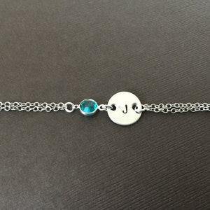 May include: A silver bracelet with a double chain design. It features a small, round, teal-colored gemstone and a circular charm engraved with the letter "J". The bracelet is displayed against a brown background.