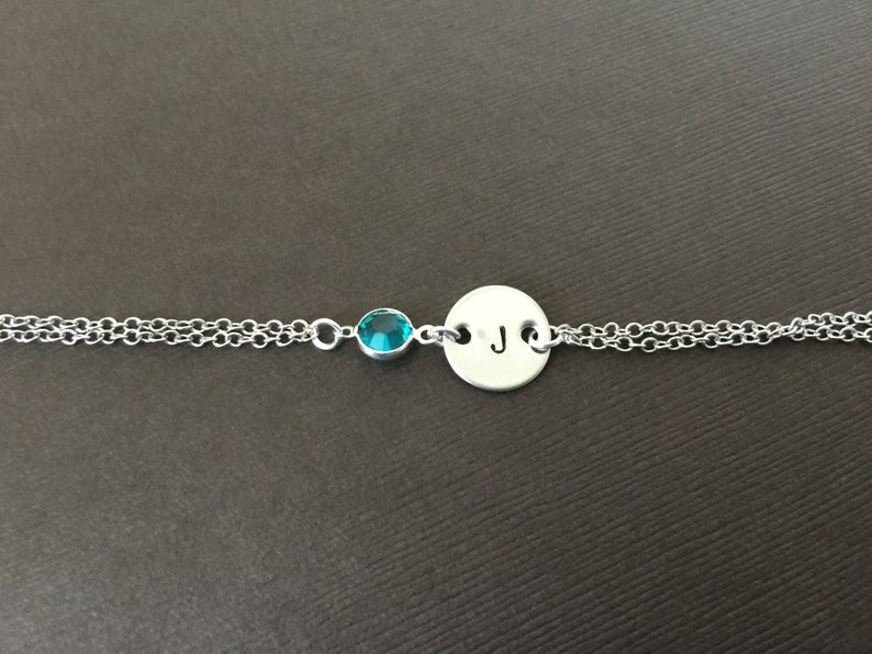 May include: A silver bracelet with a double chain design. It features a small, round, teal-colored gemstone and a circular charm engraved with the letter "J". The bracelet is displayed against a brown background.