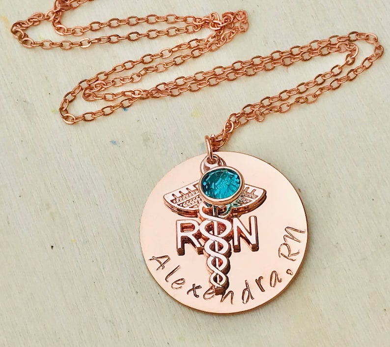 May include: Rose gold necklace with a round pendant featuring a medical caduceus symbol, the letters "RN" and the name "Alexandra RN". A small turquoise gemstone is set in the center of the pendant.