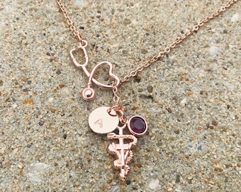 May include: Rose gold necklace featuring a stethoscope, heart, and medical symbol pendant. The necklace includes a round charm with the letter "A" and a small, round, purple gemstone. The chain and pendants are rose gold.