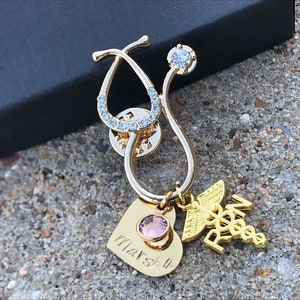 May include: Gold stethoscope pin with a pink heart charm, a clear rhinestone, and a gold charm with the letters "RN" and a caduceus. The heart charm has the inscription "Marsh 4".