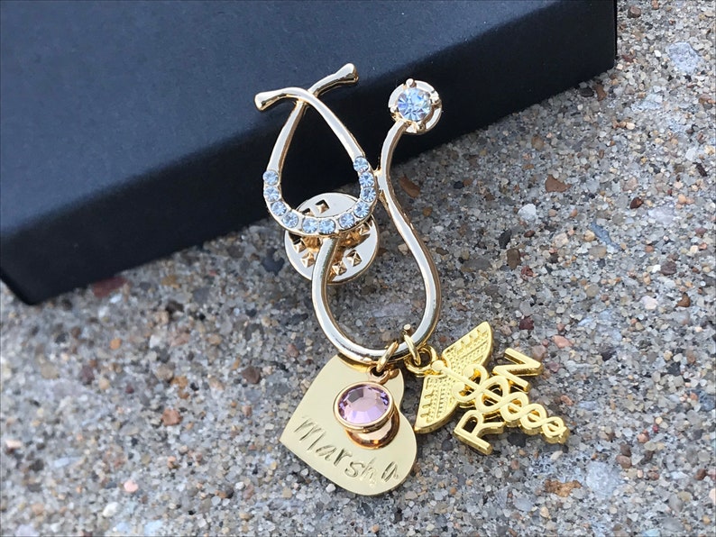 May include: Gold stethoscope pin with a pink heart charm, a clear rhinestone, and a gold charm with the letters "RN" and a caduceus. The heart charm has the inscription "Marsh 4".
