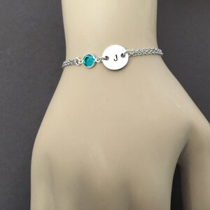 May include: A silver bracelet with a teal-colored gemstone and a circular charm engraved with the letter "J". The bracelet is on a double chain and is displayed on a mannequin hand.