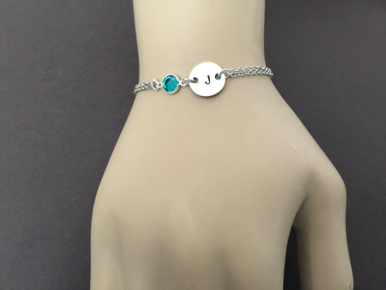 May include: A silver bracelet with a teal-colored gemstone and a circular charm engraved with the letter "J". The bracelet is on a double chain and is displayed on a mannequin hand.