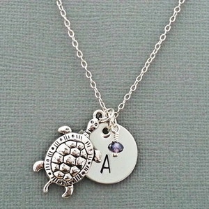 May include: Silver necklace with a turtle charm and a round charm with the letter 'A' on it. A small purple bead hangs from the charm.