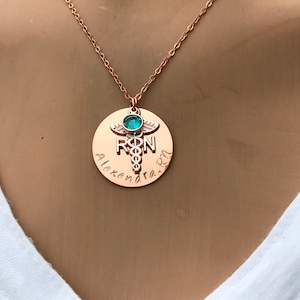 May include: Rose gold necklace with a round pendant featuring a medical caduceus symbol, the letters "RN" and the name "Alexandra" engraved on it. A turquoise gemstone is set in the center of the caduceus.