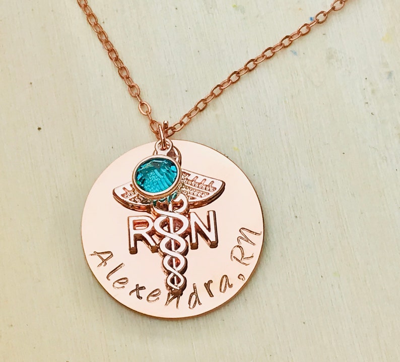 May include: Rose gold necklace with a round pendant featuring a medical caduceus symbol, the letters "RN", and the name "Alexandra RN". A small turquoise gemstone is attached to the top of the pendant.