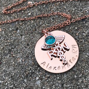 May include: Rose gold necklace with a round pendant featuring a medical caduceus symbol, the letters "RN" and the name "Alexandra, RN". A small turquoise gemstone is attached to the pendant.