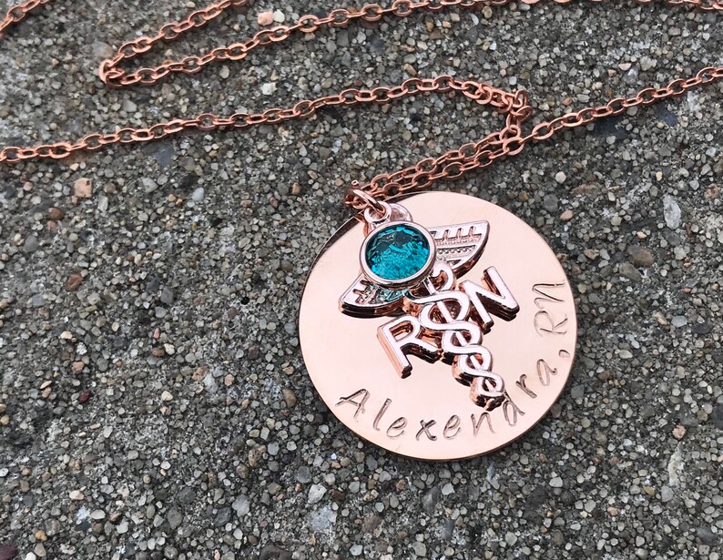 May include: Rose gold necklace with a round pendant featuring a medical caduceus symbol, the letters "RN" and the name "Alexandra, RN". A small turquoise gemstone is attached to the pendant.