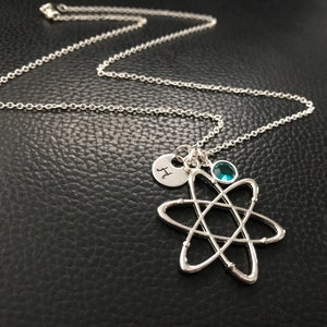 May include: Silver-toned necklace featuring an atom pendant, a small charm with the letter "H", and a teal-colored gemstone. The chain is delicate, and the atom pendant is the focal point of the necklace.