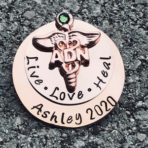 May include: Rose gold tone pendant with a green gem, a caduceus symbol, and the text "ADN" in the center. The pendant is engraved with the text "Live. Love. Heal. Ashley 2020".