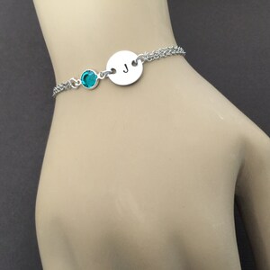 May include: A silver bracelet featuring a teal-colored gemstone and a circular charm with the letter "J". The bracelet has a delicate chain design, perfect for everyday wear. The bracelet is shown on a person's wrist.