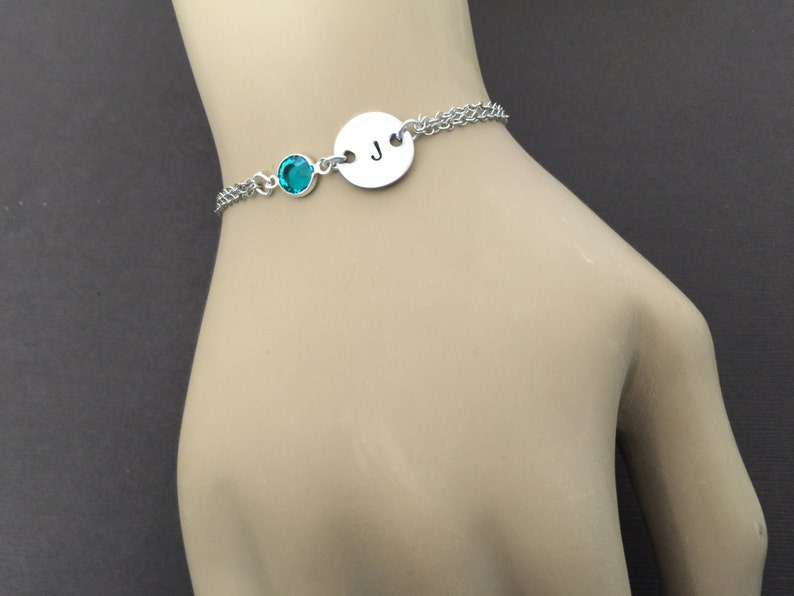 May include: A silver bracelet featuring a teal-colored gemstone and a circular charm with the letter "J". The bracelet has a delicate chain design, perfect for everyday wear. The bracelet is shown on a person's wrist.