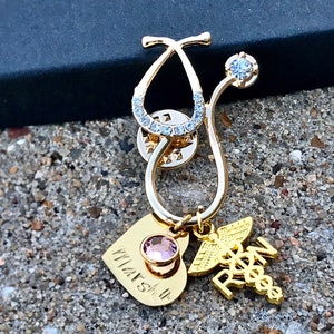 May include: Gold stethoscope charm with a pink crystal heart charm and a gold 'RN' charm. The stethoscope charm is decorated with clear rhinestones.