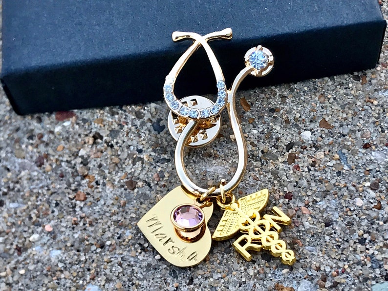 May include: Gold stethoscope charm with a pink crystal heart charm and a gold 'RN' charm. The stethoscope charm is decorated with clear rhinestones.