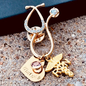 May include: Gold stethoscope pin with a clear rhinestone accent, a heart charm with a pink rhinestone, and a medical symbol charm with the letters "RN" on it. The heart charm has the word "Marsh" engraved on it.