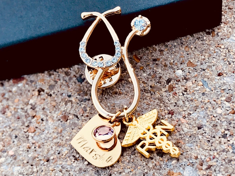 May include: Gold stethoscope pin with a clear rhinestone accent, a heart charm with a pink rhinestone, and a medical symbol charm with the letters "RN" on it. The heart charm has the word "Marsh" engraved on it.