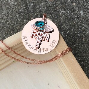 May include: Rose gold necklace with a round pendant featuring a medical caduceus symbol, the letters "RN" and the name "Alexandra. RN". A small turquoise gemstone is set in the top of the pendant.