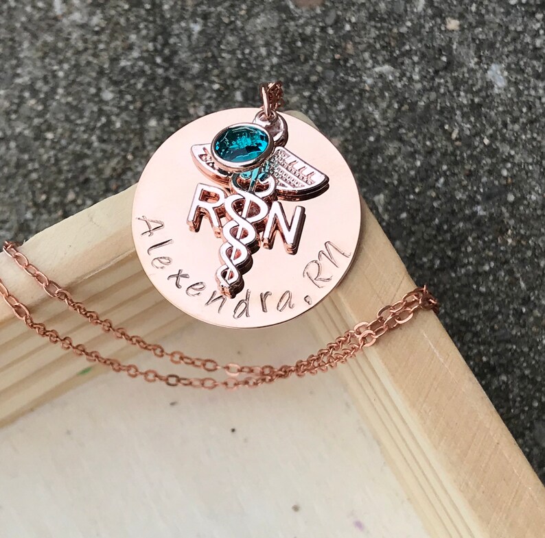 May include: Rose gold necklace with a round pendant featuring a medical caduceus symbol, the letters "RN" and the name "Alexandra. RN". A small turquoise gemstone is set in the top of the pendant.
