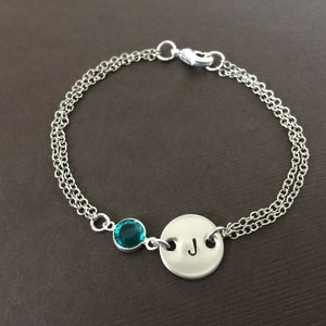 May include: Silver-toned bracelet featuring a teal-colored gemstone and a circular charm with the letter "J". The bracelet has a double chain design and a clasp closure. A personalized piece of jewelry.