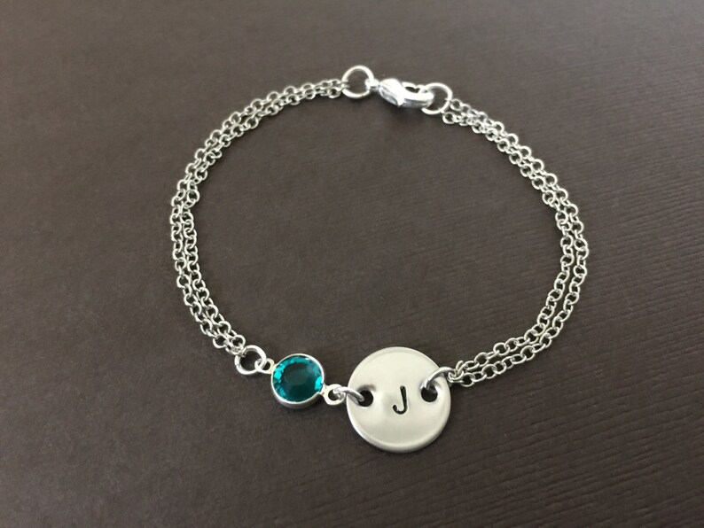 May include: Silver-toned bracelet featuring a teal-colored gemstone and a circular charm with the letter "J". The bracelet has a double chain design and a clasp closure. A personalized piece of jewelry.