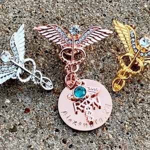 May include: Three gold, silver, and rose gold medical caduceus pins with rhinestones. The rose gold pin has a round charm with a turquoise stone and the text "RN" and "Alexandra".
