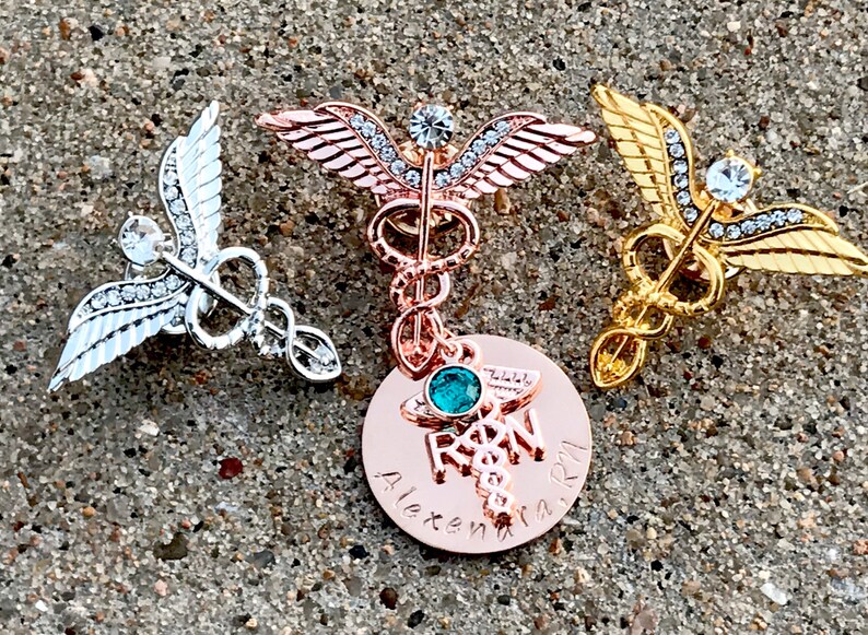 May include: Three gold, silver, and rose gold medical caduceus pins with rhinestones. The rose gold pin has a round charm with a turquoise stone and the text "RN" and "Alexandra".