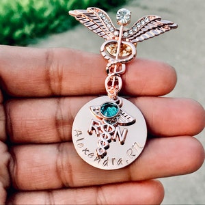May include: Rose gold medical caduceus pin with a clear crystal and a round charm with the text "Alexandra RN".
