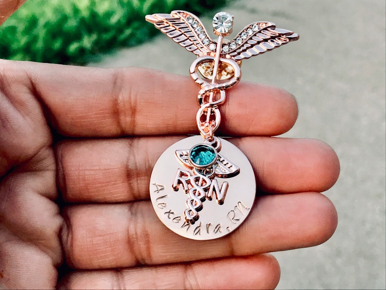 May include: Rose gold medical caduceus pin with a clear crystal and a round charm with the text "Alexandra RN".