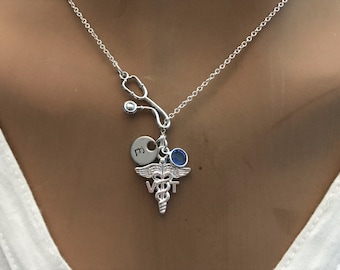 May include: Silver necklace with a stethoscope charm, a small blue stone, and a round charm with the letter 'E' on it. The necklace also features a silver medical caduceus charm with the letters 'VET' on it.