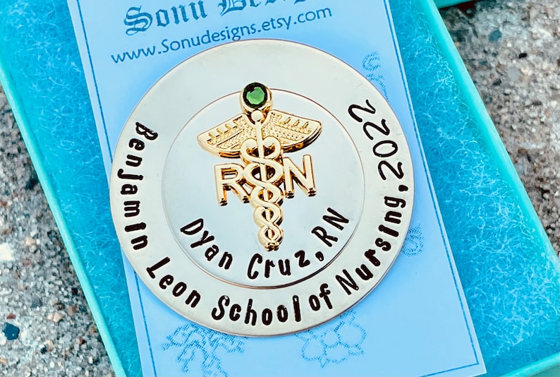 Peut inclure: Une &eacute;pingle ronde argent&eacute;e avec un symbole m&eacute;dical dor&eacute; et le texte "Benjamin Leon School of Nursing, 2022, Dylan Cruz, RN".