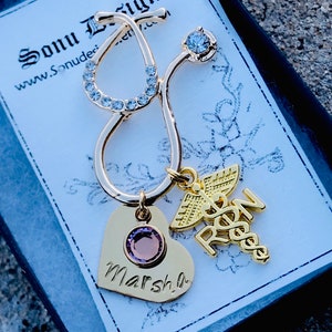 May include: Gold-tone stethoscope charm with a heart charm engraved with the name "Marsha" and a charm with the letters "RN" and a caduceus symbol. The charms are on a black background.