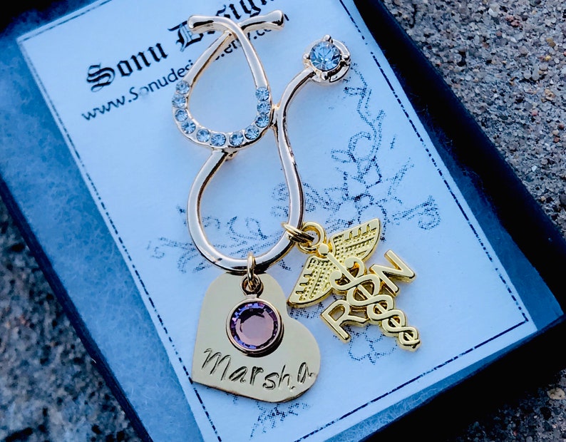 May include: Gold-tone stethoscope charm with a heart charm engraved with the name "Marsha" and a charm with the letters "RN" and a caduceus symbol. The charms are on a black background.