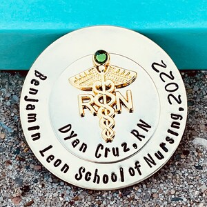 Peut inclure: Une &eacute;pingle ronde dor&eacute;e avec un bijou vert et les mots "Benjamin Leon School of Nursing 2022" et "Dyan Cruz, RN" autour d'un symbole de caduc&eacute;e.