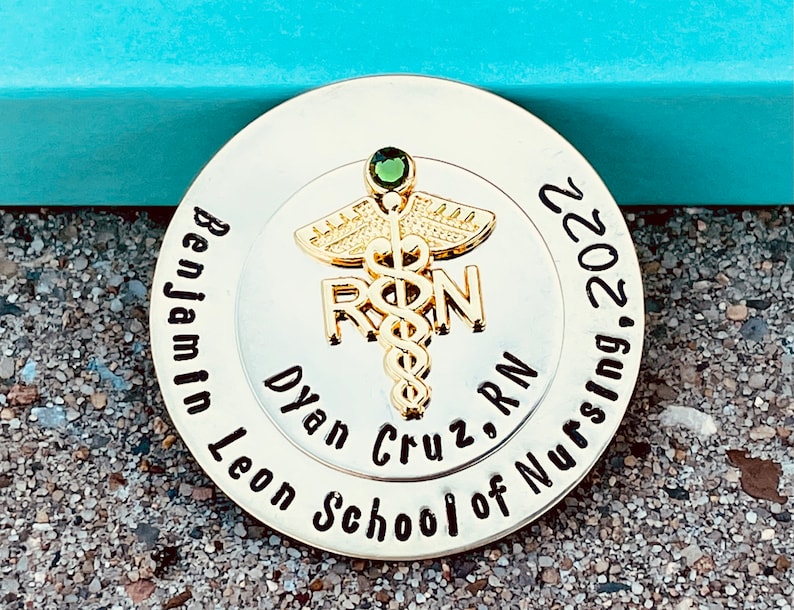 Peut inclure: Une &eacute;pingle ronde dor&eacute;e avec un bijou vert et les mots "Benjamin Leon School of Nursing 2022" et "Dyan Cruz, RN" autour d'un symbole de caduc&eacute;e.