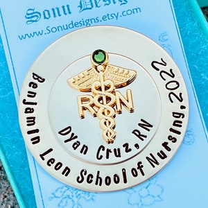 Peut inclure: Une &eacute;pingle ronde dor&eacute;e avec un bijou vert au centre. L'&eacute;pingle repr&eacute;sente un caduc&eacute;e avec les lettres "RN" et le texte "Benjamin Leon Cruz, RN School of Nursing, 2022".