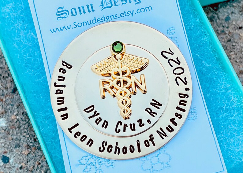 Peut inclure: Une &eacute;pingle ronde dor&eacute;e avec un bijou vert au centre. L'&eacute;pingle repr&eacute;sente un caduc&eacute;e avec les lettres "RN" et le texte "Benjamin Leon Cruz, RN School of Nursing, 2022".