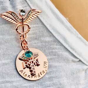 May include: Rose gold medical caduceus pin with a clear crystal and green gem. The pin has a round charm with the words "RN" and "Alexandra" engraved on it.